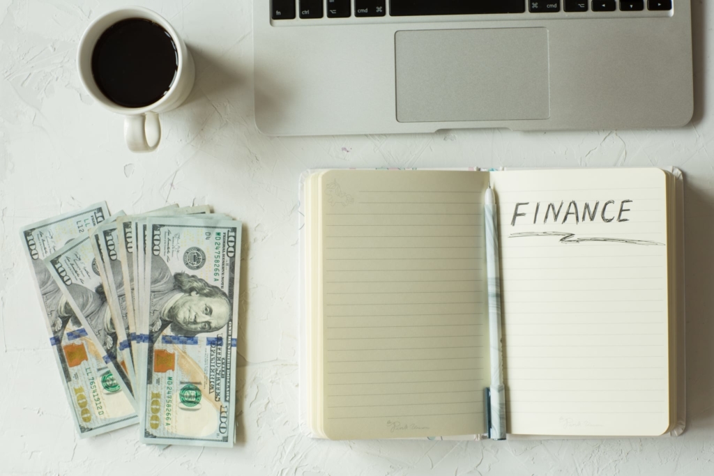 Money, notebook with 'finance' written on page, coffee, and laptop on a desk