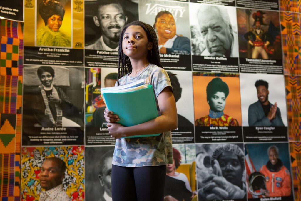 A young black female student standing holding a book with civil rights posters in the background
