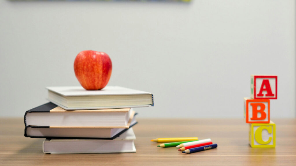 An apple on top of books, on a table with colored pencils in the middle, and colored blocks spelling ABC on the right