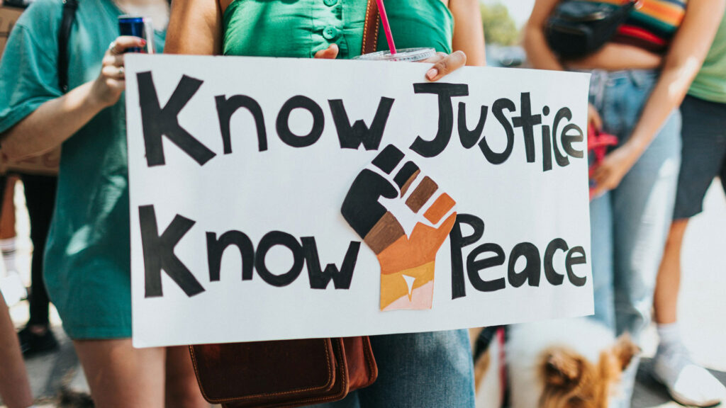 Sign held by a female protester: Know Justice Know Peace with a graphic of a closed fist in the middle
