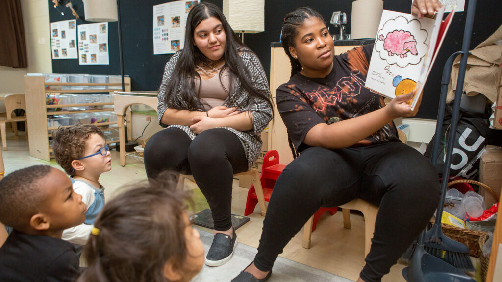 Two female student teachers read a book to preschool students seated on the floor