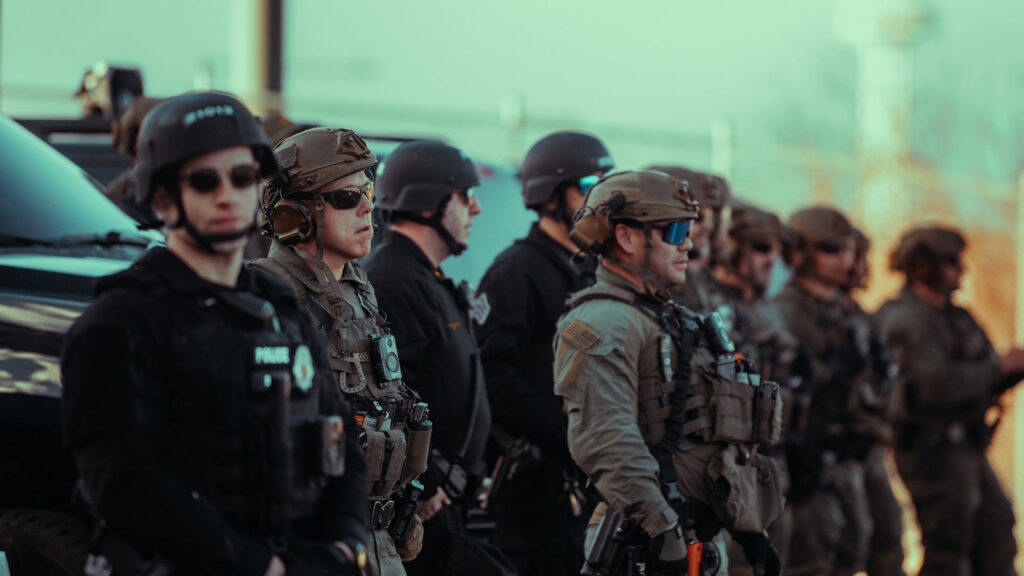 A line of armed policemen in helmets and body armor wearing sunglasses