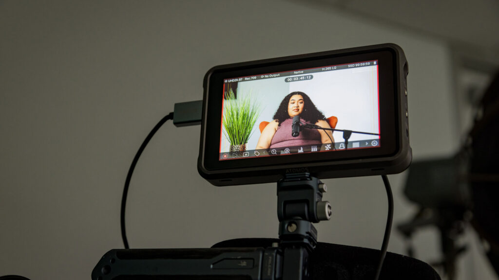 A shot of a camera screen with a female speaking to a microphone with wires coming out and placed on top of a camera stand