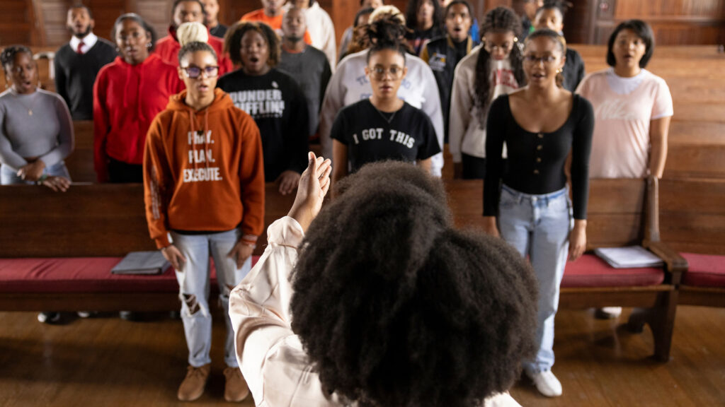 Black female leading a choir of Black students standing in pews in a college chapel