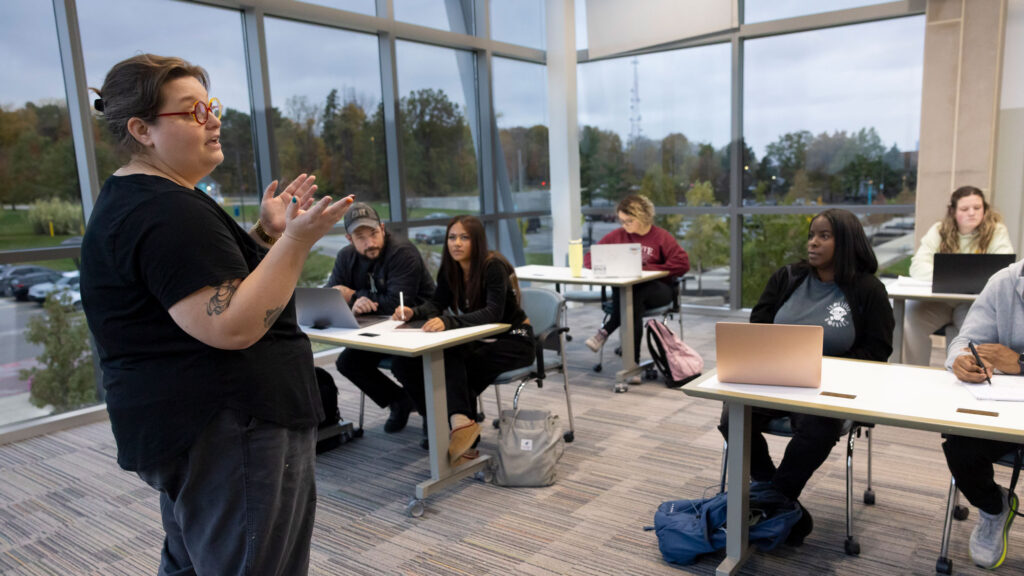 Instructor lecturing during an evening class in a windowed room overlooking a parking lot on a college campus